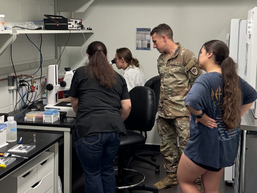 Emily working at a microscope.  Islyn standing near the microscope.  With two students observing.